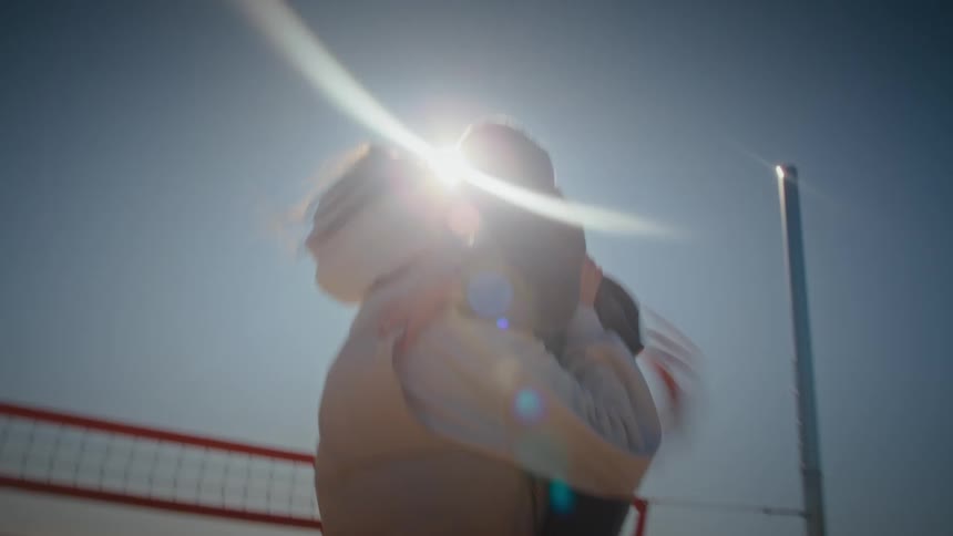 A mother lifts her child into a warm hug on a sunny beach volleyball court with bright sunlight shining overhead and the ocean in the background