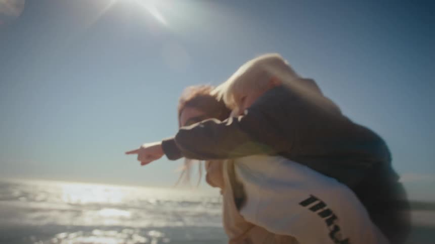 A woman wearing sunglasses and a vest gives a young child a piggyback ride while running along a sandy beach with crashing waves under a bright sun and clear blue sky