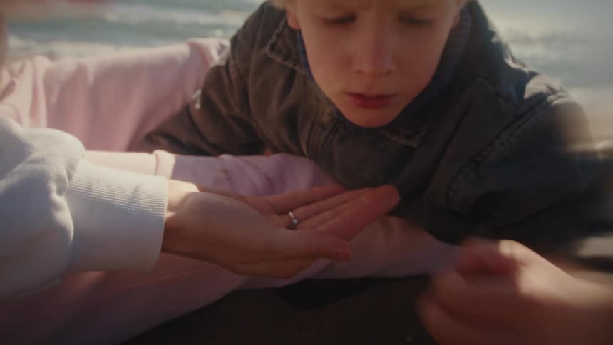 A child in a grey jacket looks closely at small objects held in the open palm of a person wearing a white sweatshirt and pink pants while they sit together on a sandy beach during a sunny day