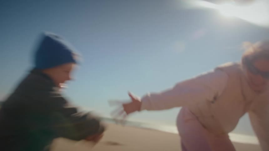 A young boy wearing a blue beanie runs across a sandy beach toward a woman standing near a volleyball netthe woman catches him in an embrace as they play together under a bright sunny sky