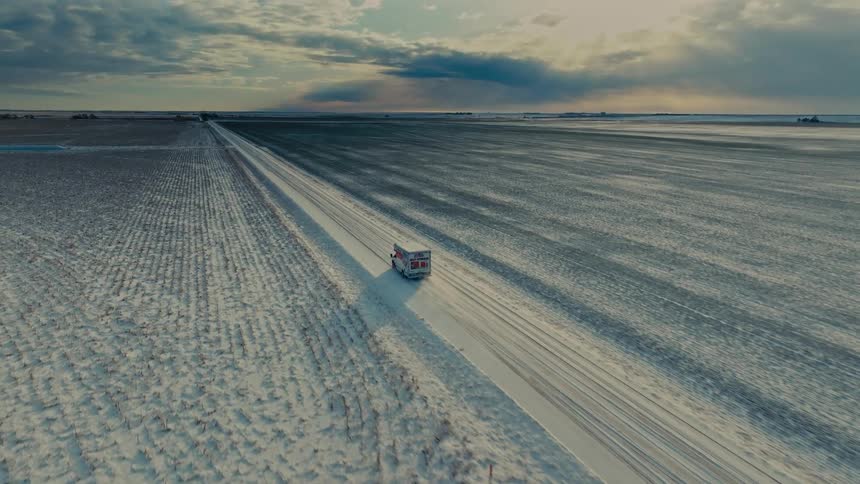 An aerial shot follows a vehicle traveling down a straight snowcovered road through vast flat farmlands the scene is set under a dramatic sky with sunlight breaking through the clouds at the horizon