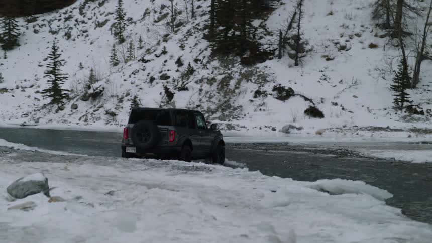 Dark suv drives through shallow icy river in snowy mountain landscape