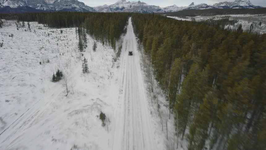 Vehicle traveling on a snowcovered road between forest with mountains in the background