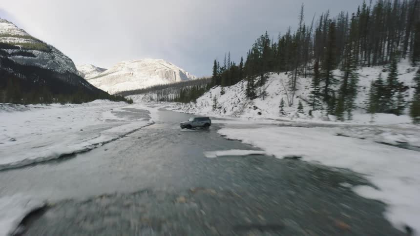 A grey off road vehicle drives across a shallow river surrounded by ice and snow in a wide mountain valley with evergreen trees under a cloudy sky