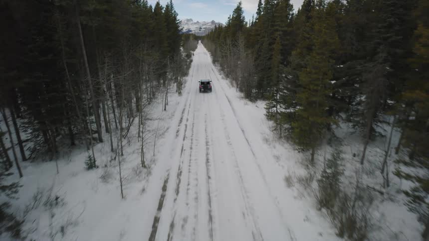 An aerial view shows a dark vehicle driving down a straight snowcovered road lined with dense evergreen forestsas the camera pulls back the road stretches toward a distant range of rugged snowcapped mountains under an overcast sky