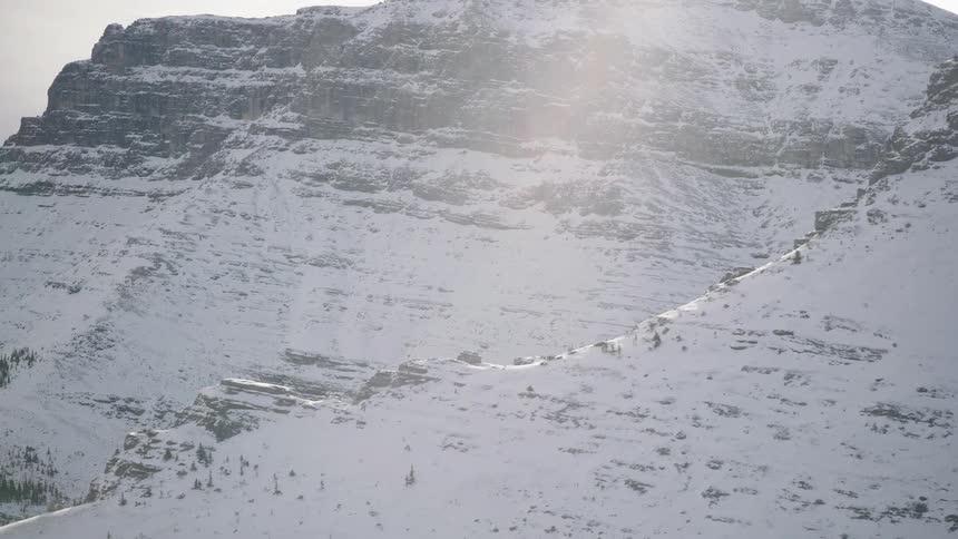 Snow covers the rough terrain of mountains as sunlight shines through a cloudy winter sky