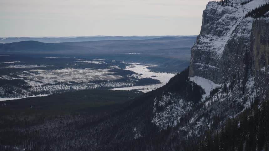 A view from a high mountain shows a steep rocky cliff and a dark forest of pine trees overlooking a vast valley with a winding frozen river under an overcast sky