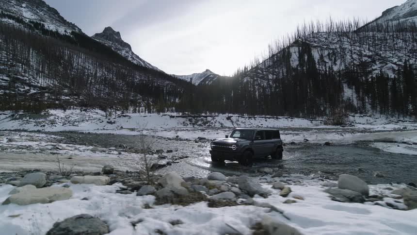 A grey suv drives through a shallow rocky riverbed surrounded by snowcovered mountains and sparse evergreen treesthe vehicle moves slowly through the water toward a snowdusted bank under a bright winter sun
