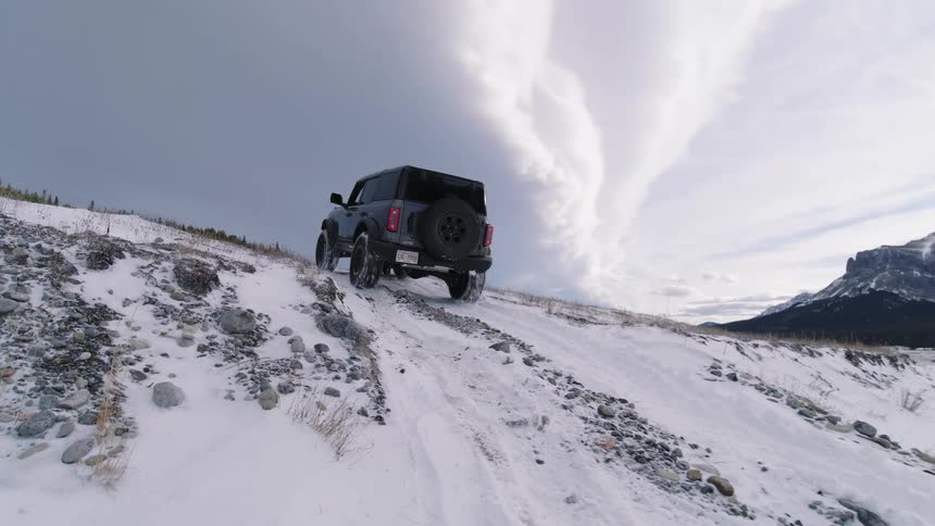 A dark grey suv drives up a snowy rocky incline toward a ridge the vehicle reaches the top of the slope against a backdrop of dramatic clouds and distant mountains