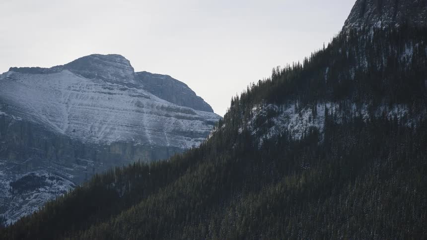 A view of a steep mountain slope covered in dark green pine trees with patches of snow under a pale sky with another rocky snow covered peak visible in the distance