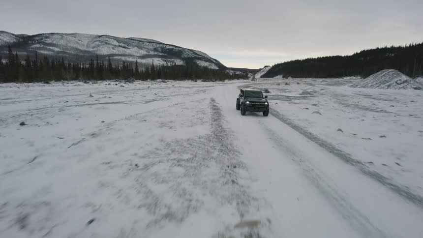 A black suv drives across a vast snow covered valley floor surrounded by distant pine forests and rugged mountains under a gray winter sky as the camera follows the vehicle