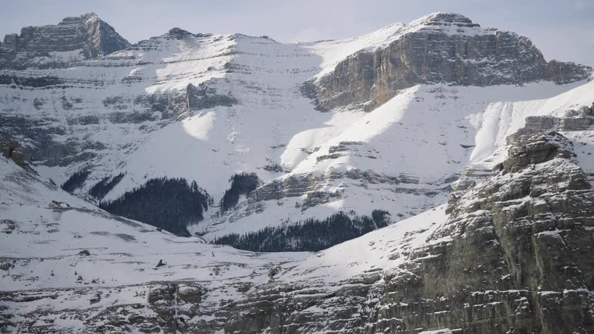 A view of a steep mountain slope covered in dark green pine trees with patches of snow under a pale sky with another rocky snow covered peak visible in the distance