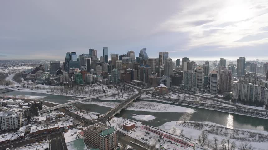 An aerial view shows a dense city skyline situated along a winding river snow covers the ground and rooftops throughout the urban landscape and along the riverbanks