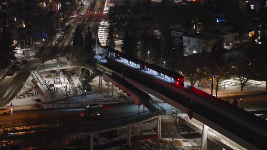 A train glides across a concrete bridge over a dark river at night its path marked by glowing red tail lightssnow dusts the surrounding urban landscape where city lights and street lamps illuminate the streets and buildings below