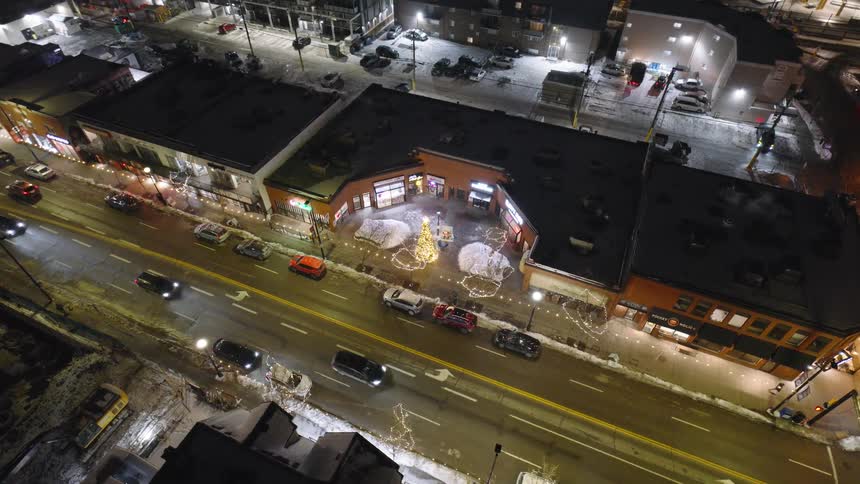 A high angle view shows cars driving along a city street at night past buildings with glowing windows and a bright holiday tree in a small plaza surrounded by snow