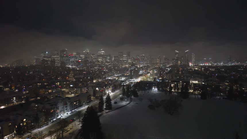 Vehicles drive past residential buildings and a park a distance from downtown on a cloudy winter night