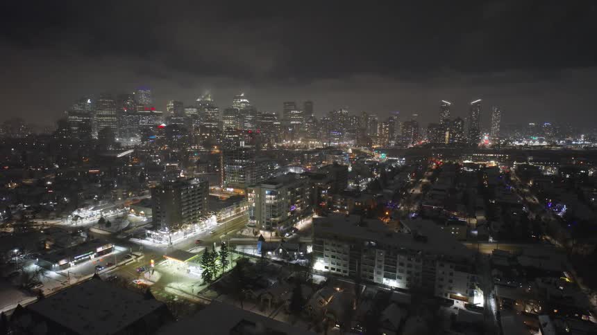 A wide view of a city at night with glowing lights from office buildings and apartment blocks under a dark cloudy sky while snow covers the ground and rooftops