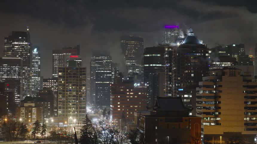 The city skyline is illuminated by a dense cluster of glowing windows and vibrant signs under a dark hazy night skysteam rises from rooftops into the cold air while the streets below are visible through a layer of snow and winter trees
