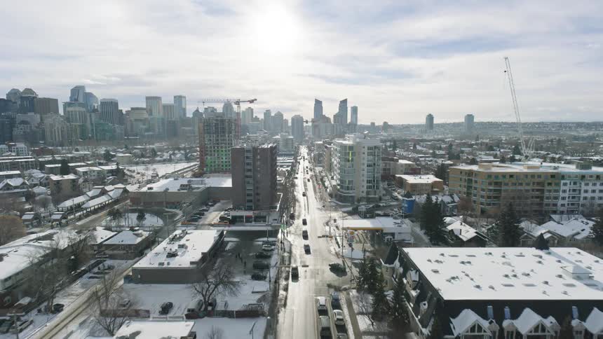 This aerial video captures a long straight road leading directly toward a distant city skyline under a bright winter sunthe surrounding landscape is a mix of modern midrise apartments and older residential buildings all draped in a thick layer of