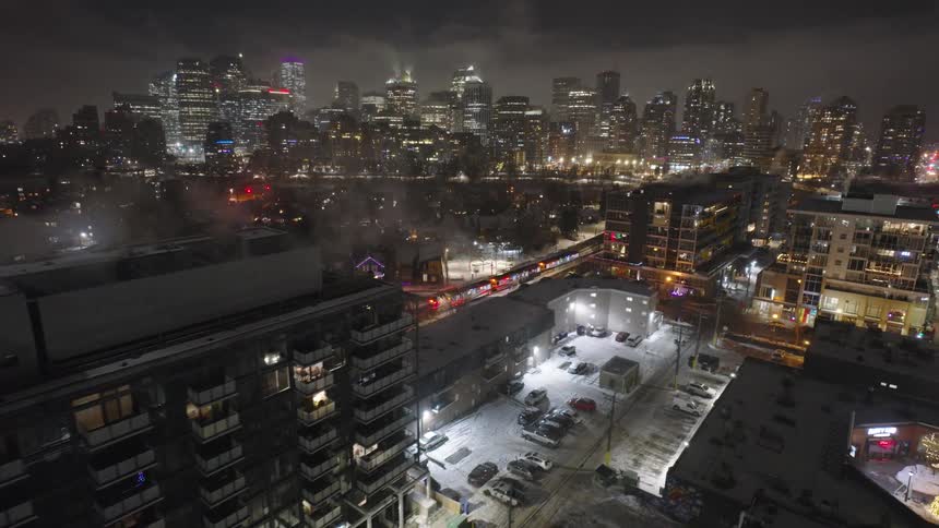 A high angle view shows a train passing through a snowy city at night with glowing lights from office towers and residential buildings under a dark cloudy sky near a river and bridge