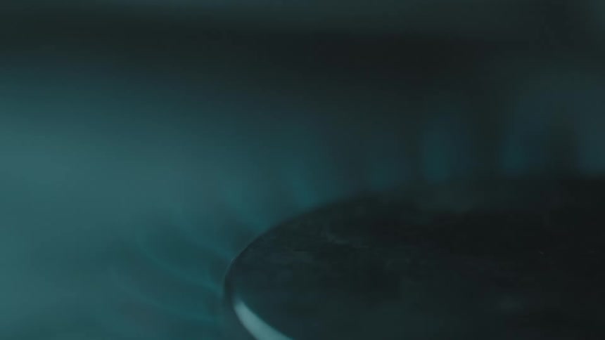 A close view shows orange sparks and small flames flickering as a blue gas fire ignites under a circular black burner on a metal stove top in a dark room