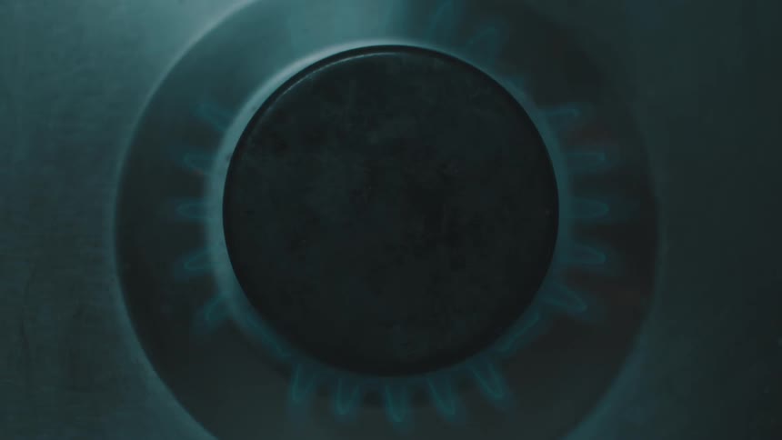 A blue flame ignites around a circular black burner on a stainless steel gas stove top in a kitchen setting