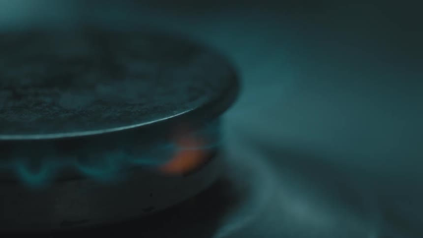 A closeup of a gas stove burner being ignited a blue flame quickly circles the burner and continues to flicker in a dim moody setting