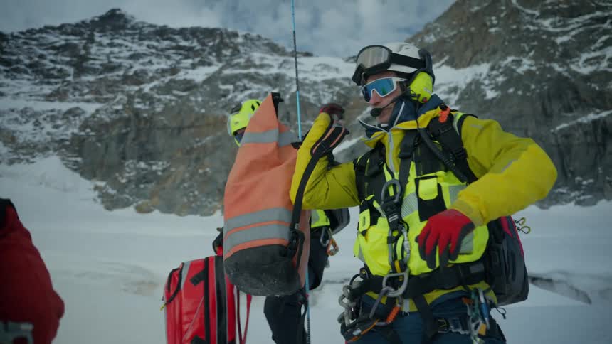 Mountain climbers pick up and carry backpacks on snowy terrain near the mountains on a cloudy day