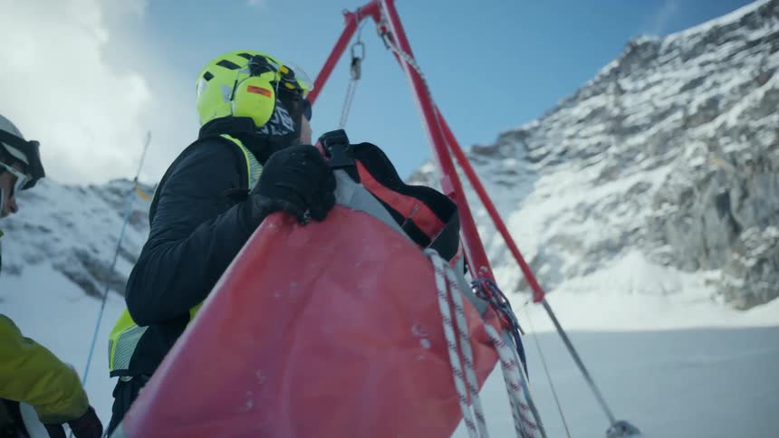 A rescue worker in a yellow safety vest and helmet operates a red lifting tripod on a snowy mountain slope while securing an orange transport bag near a rocky cliff under a clear blue sky