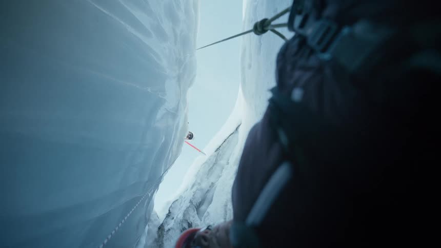 Man looking down at another man hanging from a rope on the side of a snow covered mountain cliff