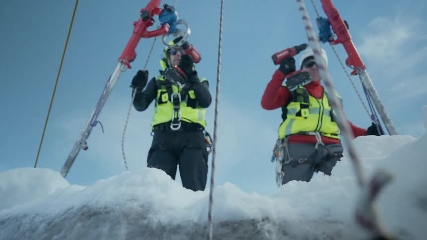 Two professionals wearing helmets and safety vests operate instruments suspended by cables over a frozen surface