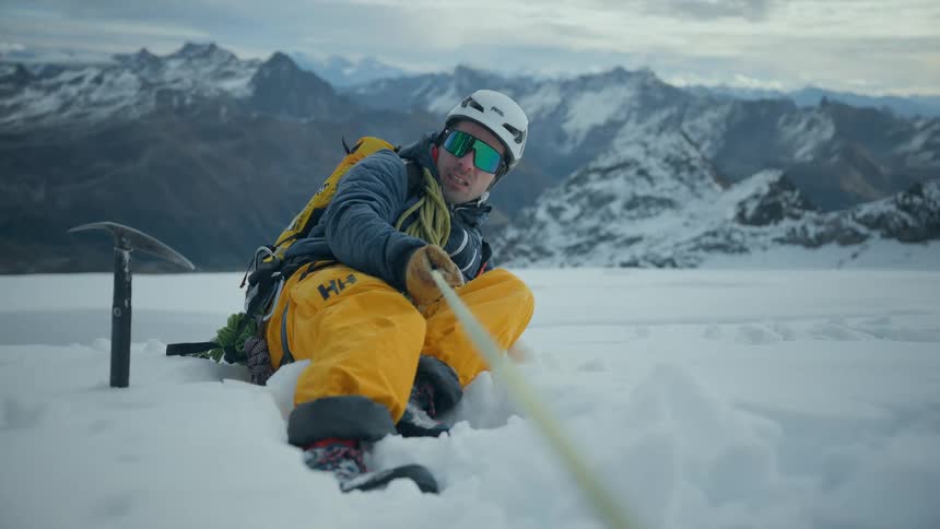 Mountain climber holding a rope sits on the snow next to an ice axe on a winter day