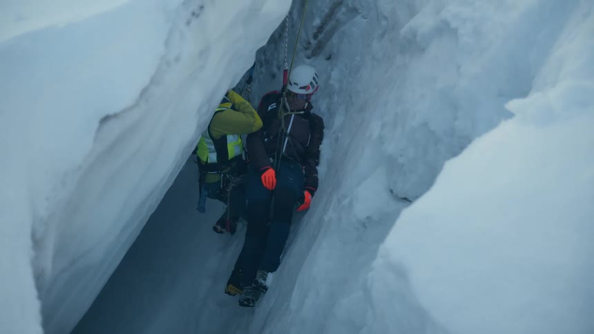 A rescue worker wearing helmet and safety gear use a rope system to carefully lift an injured person through a narrow and deep ice crevasse in a snowy mountain landscape
