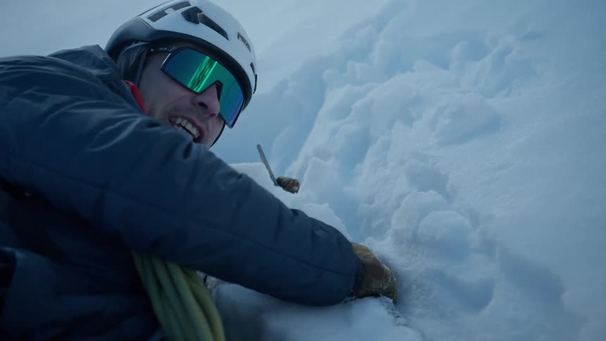 A male climber wearing a helmet and goggles grips a steep snowy mountain face while looking toward the camera