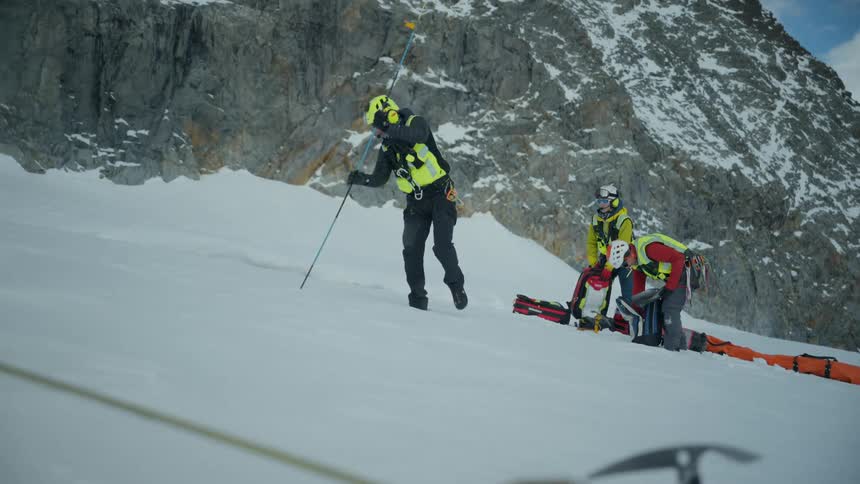 A team of rescue workers wearing helmets and winter gear prepare a stretcher on a snowy mountain slope while another worker uses a long probe in the snow during a bright day
