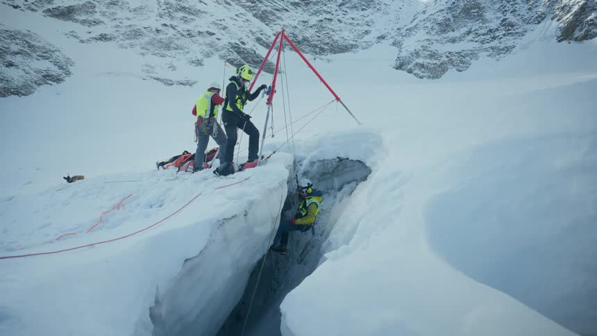 A team of rescue workers in helmets and winter gear uses a red tripod and ropes to lower a person into a deep ice crevice on a snowy mountain slope under a bright sky