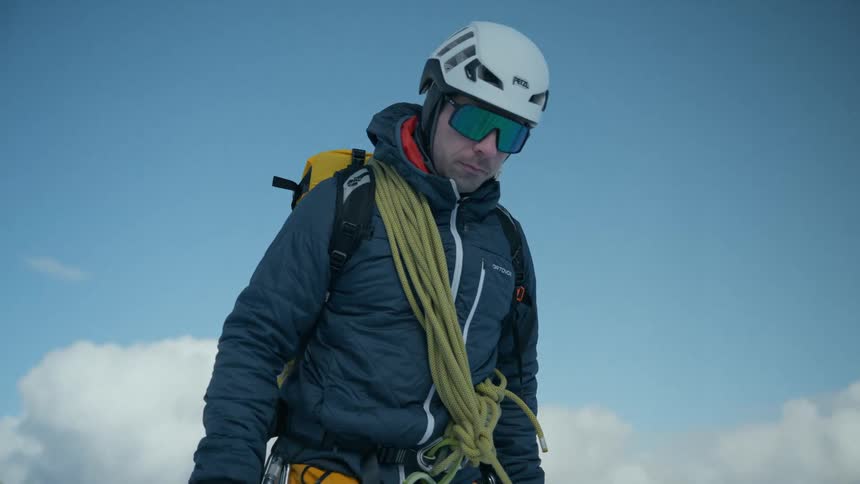 A mountain climber wearing a white helmet and orange pants walks through deep snow on a mountain ridgehe carries a coiled rope over his shoulder and an ice axe in his hand while a vast mountain range stretches out behind him