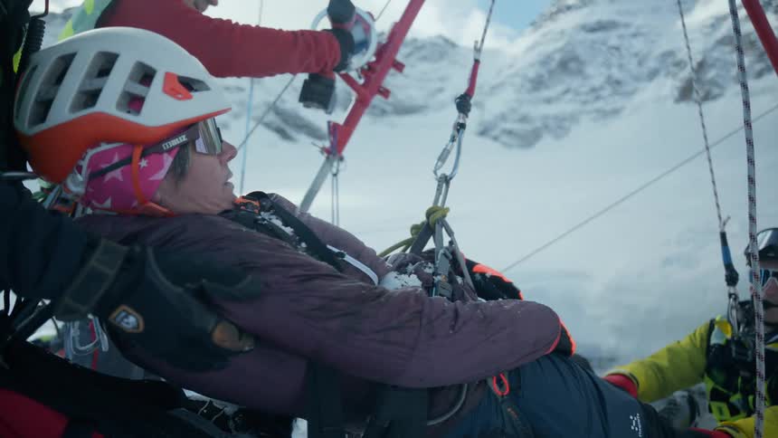A rescue team secures an injured woman in a harness on a snowy mountain as they prepare for a winch evacuation while a medic monitors her condition near heavy equipment against a steep alpine backdrop during a bright day in the cold snow