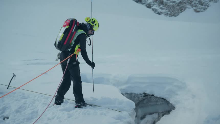 One mountain climber sits and holds a rope while another attached to a separate rope probes the snow