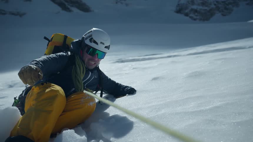 A mountaineer wearing yellow pants and a helmet slides down a steep snowy mountain slope while holding a tensioned rope and an ice axe during a bright day