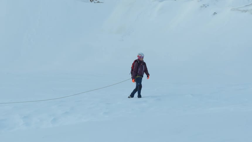 Person wearing helmet and backpack walks across a snowcovered surface pulling a rope