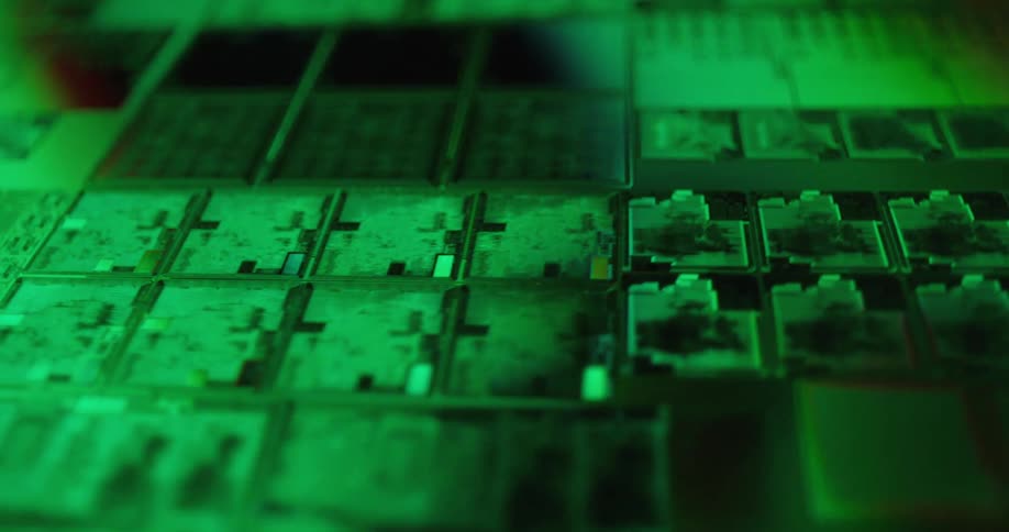 A close view shows rows of green computer microchips with small gold components glowing under a shifting green light in a dark technical setting