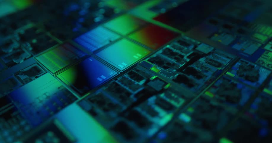 A close view shows rows of square computer microchips with intricate circuits glowing under shifting red and green and blue lights in a dark technical setting