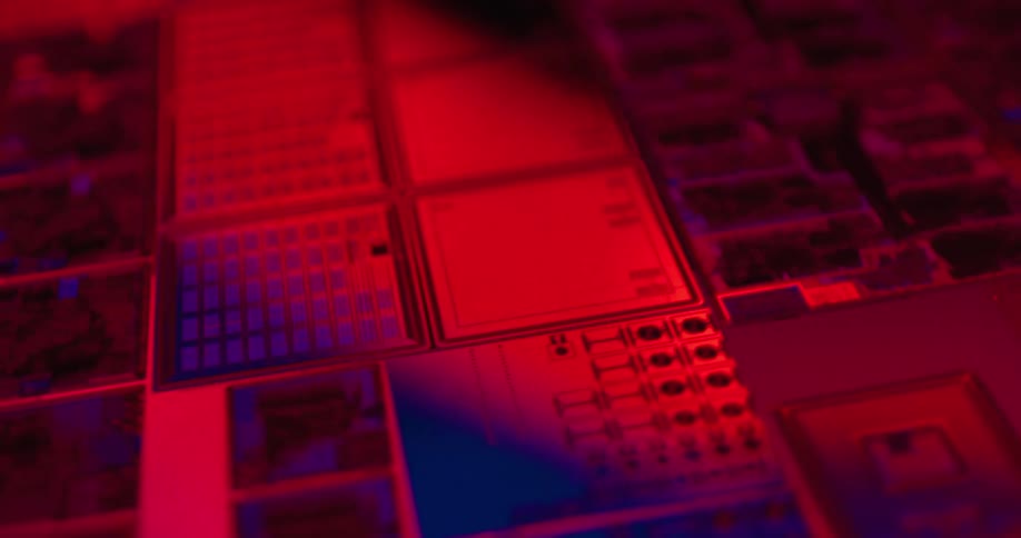 A close view shows rows of square computer microchips with intricate circuits glowing under shifting blue and red and purple lights in a dark technical setting