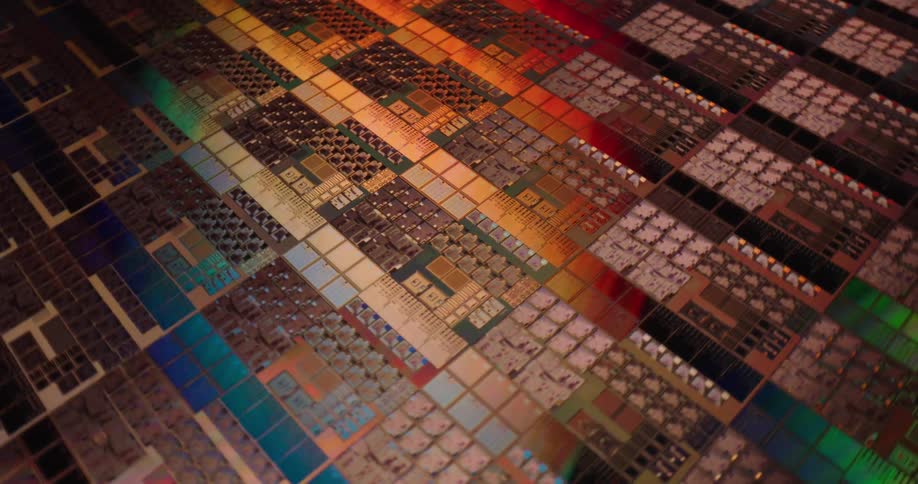 A close view shows a silicon wafer with a complex pattern of computer chips and shimmering colorful light reflections moving across the dark surface during the manufacturing process