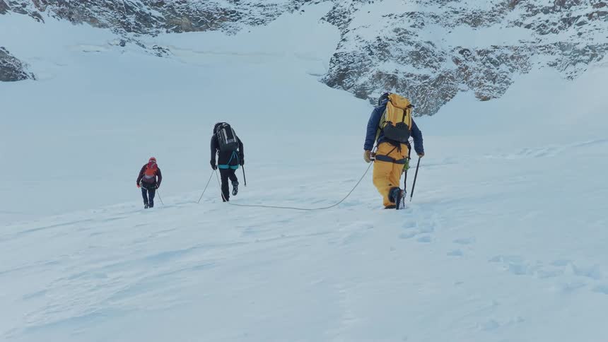 Three people equipped with backpacks and poles climb a snowcovered slope between rocky mountains