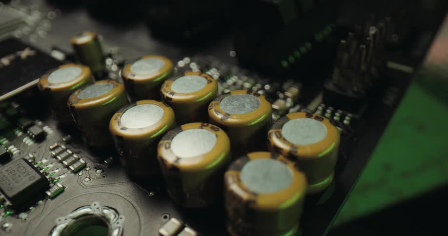 A dramatic closeup pan across a row of goldcapped capacitors on a computer motherboardthe components are highlighted by a sequence of cycling colored lightsshifting from warm amber to cool blue and neon greenagainst a dark background