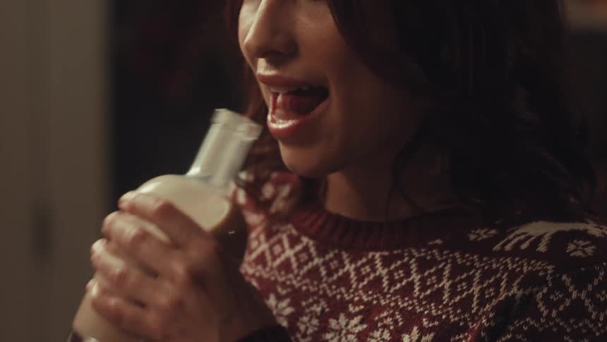 A woman wearing a festive holiday sweater licks the top of a glass bottle containing a creamy beverageshe then pulls the bottle away with a playful smile on her face