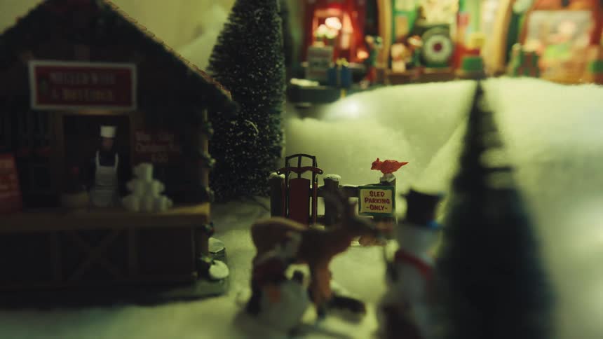A close view of a miniature holiday village scene with a small wooden hut and figurines including a reindeer and a snowman set against a snowy ground and festive lights