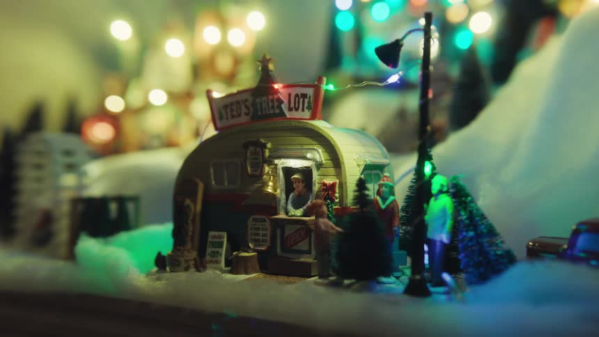 Small christmas decorative figurines sit on white as colorful christmas lights shine in the background